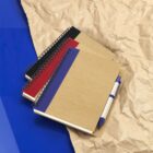 Three spiral-bound notebooks with brown covers and colored spines (black, red, blue) are stacked on crumpled brown paper, with a blue pen clipped to the top notebook. The background features a vibrant blue surface.