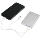 A smartphone connected to a slim, silver power bank with a white charging cable. The power bank has a USB port and an indicator light showing its in use.