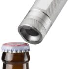 Close-up of a silver, textured bottle opener aligned with the cap of a brown glass bottle. The bottle has a white and red crowned cap, and the openers metallic surface is distinctly visible against the background.