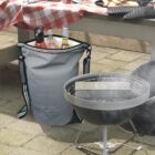 A gray cooler bag filled with colorful bottled drinks is placed next to a small grill. The grill is smoking on a patio. Nearby, a table with a red and white checkered cloth holds kitchen items.