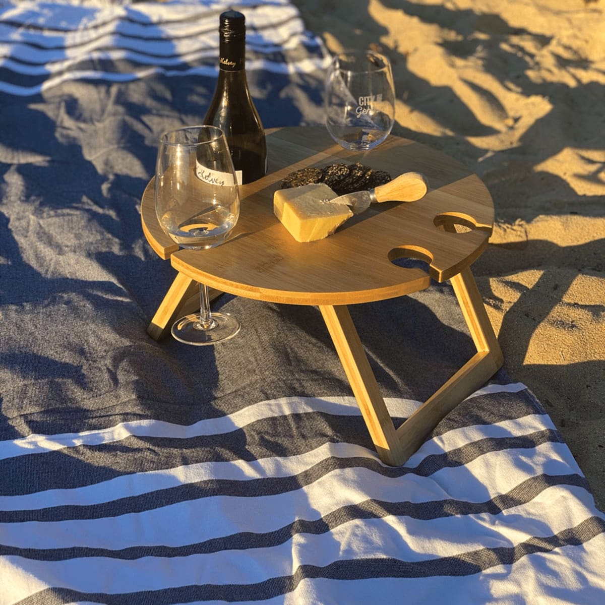 A portable wooden picnic table on a striped blanket, holding a wine bottle, two wine glasses, and a cheese block with a knife. The scene is set outdoors on a sunny day, with a sandy beach as the backdrop.