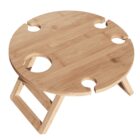 A round wooden picnic table with four recessed slots for holding wine glasses. It has foldable legs for easy transport and storage. The natural wood finish enhances its rustic appearance.