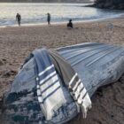 An overturned boat on a sandy beach has two striped towels draped over it. The ocean is in the background, with three people walking and sitting near the water. The scene is calm and peaceful.