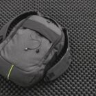 A grey backpack with a black strip and earbuds placed on it rests on a textured, dark foam surface. The backpack features a small green accent stripe and the word Zoom is visible near the bottom.