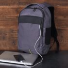 A gray backpack with a black accent rests against a wooden wall. A white charging cable extends from the backpack to a smartphone atop a closed laptop on a dark surface. The setup suggests the backpack has a built-in charging port.