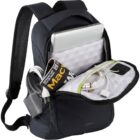 A black backpack is open, revealing a laptop, a rolled magazine, a power bank, and charging cables inside its compartments. The interior lining is white with a geometric pattern. The backpack has padded straps for carrying.