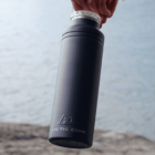 Hand holding a black Arctic Zone bottle against a blurred background of water and rocky shore. The bottle has a matte finish and a silver cap.