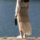 Person standing on rocky ground near a body of water, holding a black water bottle. They are wearing a white sweater, a long floral skirt, and white sneakers. The scene is serene, with rippling water visible in the background.