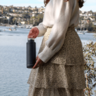 A person in a white sweater and patterned skirt holds a black insulated water bottle. They are standing by a body of water with boats and trees in the background.