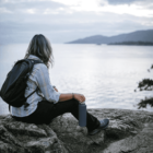 A person with long hair, wearing a plaid shirt and backpack, sits on a rock holding a bottle. They gaze at a serene body of water with distant mountains under a cloudy sky.