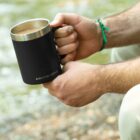 A person holding a black Arctic Zone mug with a silver rim, filled with a hot beverage. They are wearing a green and black braided bracelet, sitting outdoors on a grassy surface.