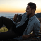 A person in a denim shirt and dark pants is sitting on a rock, smiling and holding a mug. The background features a sunset over a mountainous landscape.