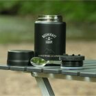 A black insulated food jar with Bushkraft Equip and Arctic Zone logos sits on a portable camping table. Next to it are a lid and a spoon. The background is blurred greenery.