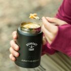 A person in a pink sweater holds a black thermos labeled Bushkraft Equip and Arctic Zone, using a spoon to eat soup or noodles from it. The background is blurred, suggesting an outdoor setting.