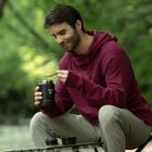 A person in a maroon hoodie is sitting on a bench outdoors, holding an open container and scooping food with a spoon. The background is green and leafy, suggesting a park or natural setting.