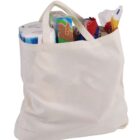 A white reusable shopping bag filled with various groceries, including toilet paper rolls, a box of pasta, a snack pack, and a loaf of bread. The bag has two handles and appears to be made of fabric.