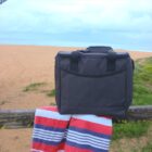 A soft cooler bag sits on a wooden fence with a striped towel draped over it. The background shows a sandy beach and a cloudy sky.