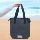 A hand holds a black tote bag on a sandy beach with the ocean in the background.