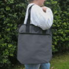 Person wearing a large gray tote bag on their shoulder, standing outdoors by green bushes.