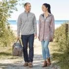 A man and woman walk along a sandy path towards a beach. The man wears a gray sweater and jeans, carrying a tote bag. The woman wears a light zip-up coat and jeans. Greenery lines the path, and the sea is visible in the background.
