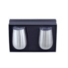 A set of two stainless steel tumblers with transparent lids, placed upright in a black foam-lined rectangular gift box.