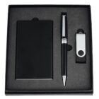 A black stationery gift set contains a notebook, a black pen with silver accents, and a silver USB drive, all neatly arranged in a black foam-lined box.