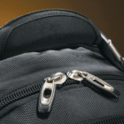 Close-up of a black fabric backpack with two metallic zipper pulls. The texture of the fabric is visible, and the zippers are slightly open, showing the top of the backpack against a warm brown background.