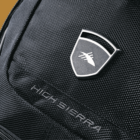 Close-up of a black High Sierra backpack showing a textured fabric with a logo featuring a stylized bird inside a shield. The brand name HIGH SIERRA is embroidered in white near the logo on the backpack.