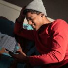 A person wearing a gray beanie and red shirt is lying on a patterned blanket, smiling while looking at their phone. They have earbuds in and sunlight is casting shadows on the floor.