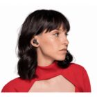 A person with shoulder-length dark hair is wearing wireless earbuds. They are looking to the side, dressed in a red top with a unique cutout design. The background is plain white.