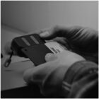 A person holding a compact device and a card marked with ZONE and 95C. The device has a loop attached, and the individual is wearing a long-sleeved, plaid shirt. The setting appears to be indoors, and the photo is in grayscale.