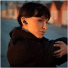A person with earbuds is stretching outdoors at sunset. They are wearing a dark jacket. Rows of houses are visible in the blurred background.