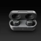 Black wireless earbuds placed inside an open charging case against a dark background. The case has a smooth finish and a small indicator light on the front. The earbuds feature a logo on their surface.