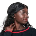 A person with long wavy hair wears wireless earbuds and looks to the side, smiling slightly. They are wearing a black sweater with red trim and have their hand gently touching their neck. The background is plain white.