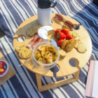 A round wooden picnic tray on a striped blanket holds strawberries, brie cheese, crackers, salami, a tub of olives, and a thermos. Sunglasses and a book are nearby, creating a cozy outdoor scene.