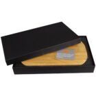 A black rectangular box partially open to reveal a wooden item inside. The item is shaped like a board and sits in a foam lining. A small card with an orange logo and text is placed on the wood.