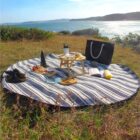 A picnic setup on a striped blanket overlooks a scenic coastal view. The blanket holds a small table with snacks, a bottle, a book, and a tote bag. Shoes are placed to the side, while the sun shines on the grassy landscape and the ocean in the background.