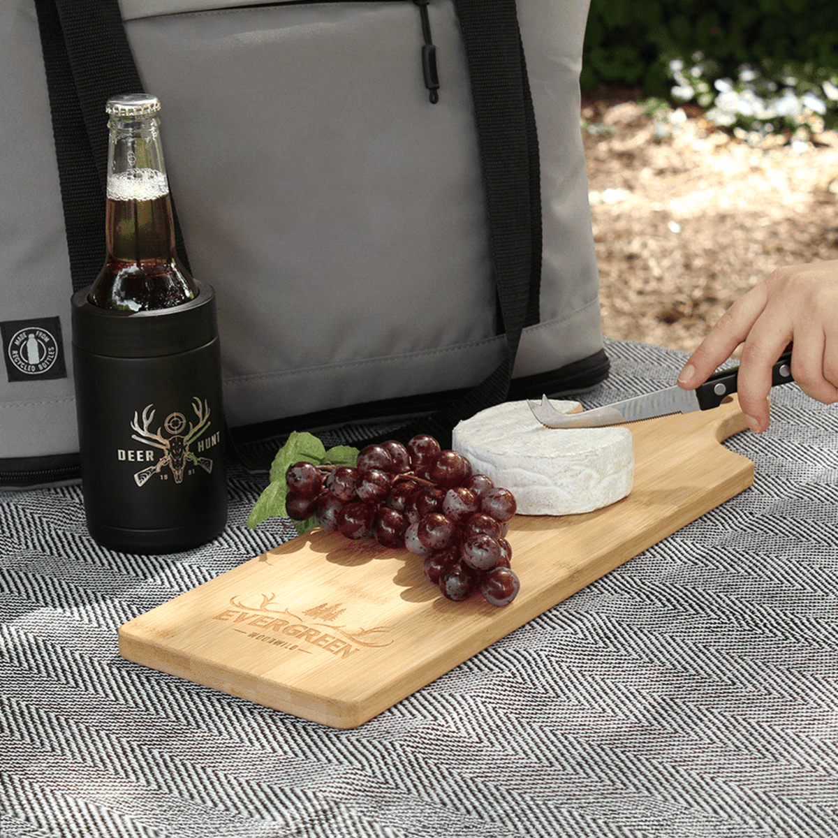 A hand slices cheese on a wooden board with red grapes. A bottle in a Deer & Hunt koozie and a gray insulated bag are nearby. The setup is on a patterned cloth outdoors.