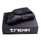 A black TREKK flashlight is placed on a TREKK-branded box. The flashlight has a textured grip and comes with a black carrying pouch.