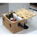 A wicker picnic basket is open on a table. It contains two bottles of wine and a soda can. A wooden lid, propped vertically, holds four wine glasses in built-in slots. A fabric strap is attached to the basket.