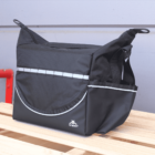A black insulated lunch bag with a zipper and front pocket, featuring a white logo and trim, sits on a wooden pallet against a gray wall.