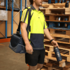 A person in a bright yellow and blue work shirt and black shorts holds a dark water bottle and carries a blue duffel bag. They stand beside stacked wooden pallets in a warehouse environment.