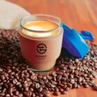 Reusable coffee cup on coffee beans with Sydney Transit logo and a blue lid.