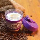 Purple travel mug with coffee and foam, surrounded by coffee beans on a wooden surface.