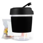 Transparent cup with black lid and sleeve, near a bag of colorful snacks.