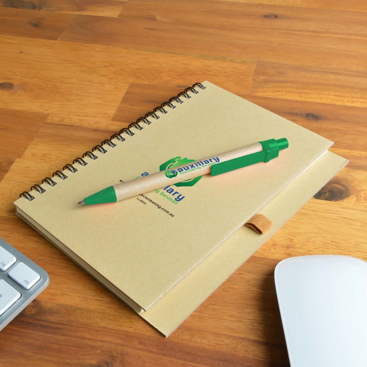 Green pen on a spiral notebook on a wooden desk with keyboard and mouse nearby.