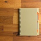 A closed spiral notebook with an orange pen on a wooden surface.
