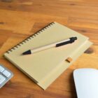 A pen on a spiral notebook with a keyboard and mouse nearby on a wooden desk.