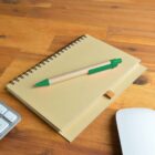 Notebook with pen on wooden desk, near a wireless keyboard and mouse.