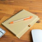 Notebook with pen on a wooden desk, next to a keyboard and a white mouse.